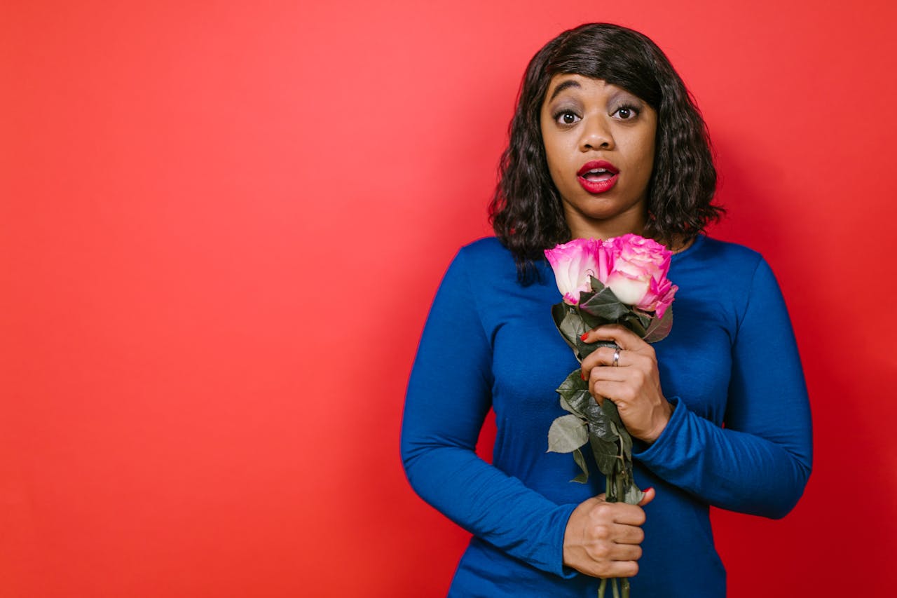 about-us A woman in blue holds pink roses with a surprised expression against a vibrant red backdrop.